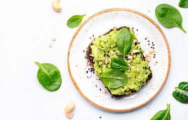 Avocado toast on rye bread with spinach leaves, cashew nuts and sesame seeds on plate, white table...