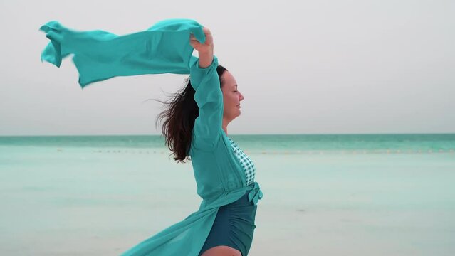 Woman in turquoise airy dress with pareo over head stands on ocean beach in windy day enjoying sea breath. Brunette female in summer holidays vacations on tropical resort. Travel, Generative AI