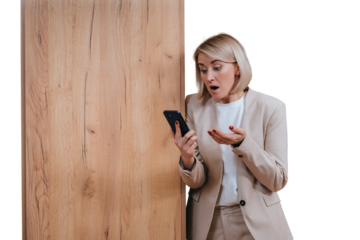 Shocked young German businesswoman in beige suit at office leans on wooden column holds phone looks at screen with wide opened eyes and mouth surprised reads message against transparent background.