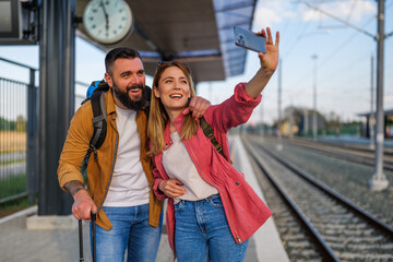 Happy couple is standing at railway station and waiting for arrival of their train. They are taking selfie.