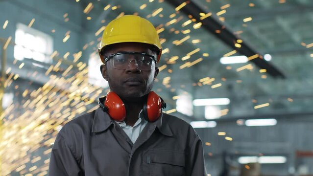 African-American worker wearing hard hat and goggles is staring intently at camera. Employee looks calm on working background of production. Ethnic diversity in construction.