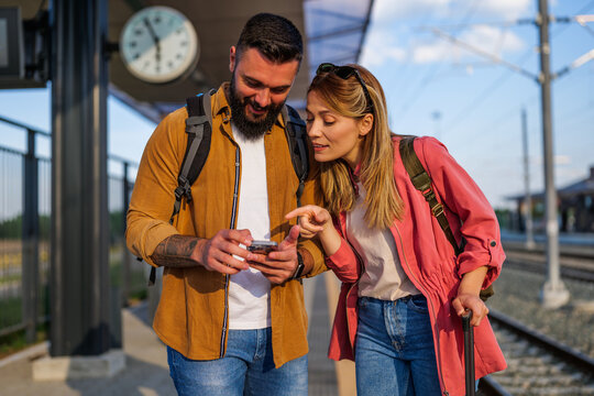 Happy couple is standing at railway station and waiting for arrival of their train. They are looking at map on smartphone.