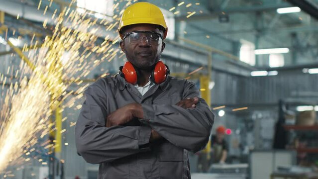African american employee with special headphones and protective helmet on working background. Manufacturing professional worker smiling and looking at camera with arms crossed.