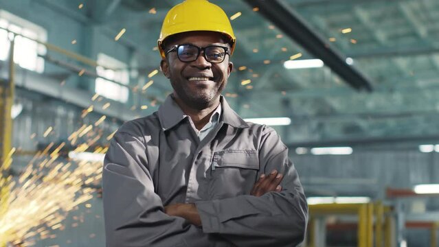 African-American employee wearing casque and glasses is smiling and looking at camera. Worker looks joyful with arms crossed. Multiplicity of production professions.