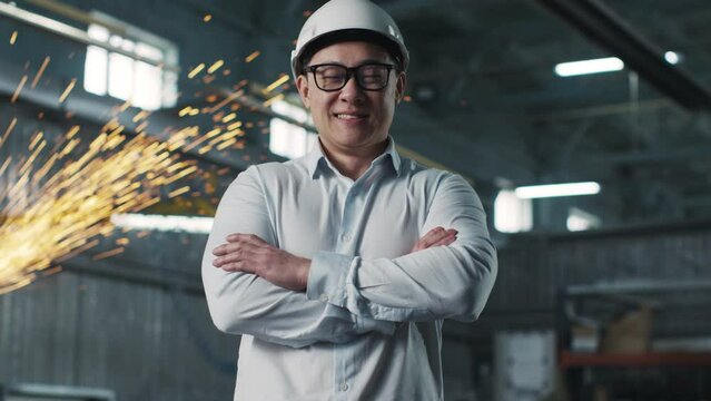 Asian male wearing white shirt, safety glasses and hard hat looks at camera. Engineer surrounded by equipment stands in background of work process with crossed armes and smiles.