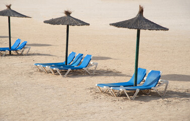 Tumbonas azules junto a sombrillas de paja en la arena blanca de una playa paradisíaca en la localidad turística de Fuerteventura en las Islas Canarias.