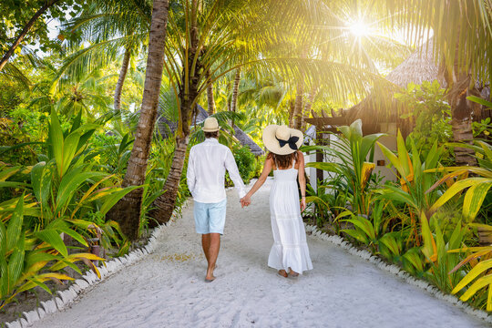 A luxury couple in white summer clothing walks on a tropical island with lush green and Palm Trees at the Maldives, Indian Ocean