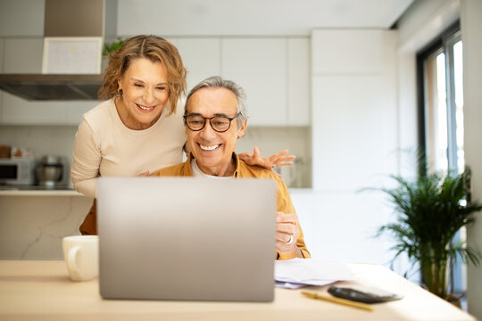 Happy senior spouses paying bills online from home, sitting in front of laptop with papers, kitchen interior, copy space Generative AI
