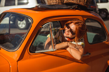 Young beautiful woman in a silk scarf and sunglasses sits behind the wheel of an orange retro car