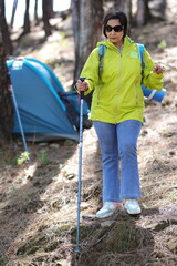 Beautiful senior women in early 60 hiking in forest, sightseeing and enjoying in nature. She s standing in front of tent.