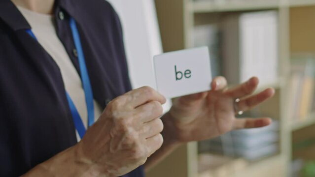 Medium close up shot of female teacher holding word card with English verb and talking to students while giving lesson in language school