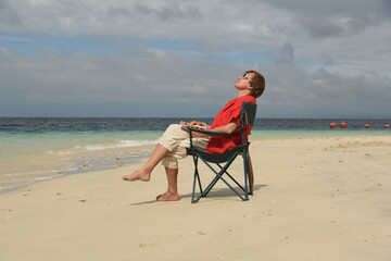 Mature woman relaxing on the beach and looking up to the sky. Senior woman wearing red shirt and sitting on the cheir. Active lifestyle after retirement. Side view