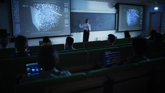 Young University Professor Explaining the Importance of Artificial Intelligence to a Group of Diverse Students in a Dark Auditorium. Female Teacher Showing Neural Network Eco System on Two Big Screens
