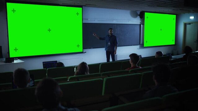Young Male Teacher Giving a Lecture, Showing Slides on a Green Screen Mock Up Display to a Diverse Multiethnic Group of Female and Male Students in Dark College Auditorium