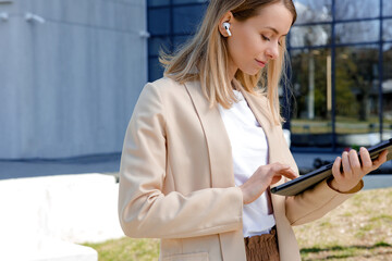Fototapeta premium Caucasian businesslady in wireless earphones using digital tablet and checking social media while standing near modern office center. Concept of people, lifestyles and gadgets