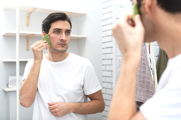 Handsome man doing skin care procedure by Scraping Facial Massage Tools such as Gouache Scraper for anti-aging Gua Sha board made by natural Jade Stone Gua Sha scraper