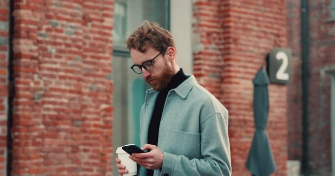 Redhead bearded guy using smartphone and holding coffee-to-go while walking. Generative AI