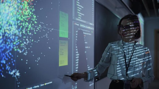 Portrait of a Young Female Professor Explaining Big Data and Artificial Intelligence Research Project in a Dark Room with a Screen Showing a Neural Network Model. Computer Science Education in College