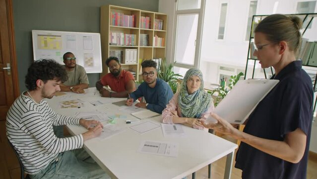 Woman holding clipboard and explaining lesson while teaching group of multi-ethnic immigrant students in language school