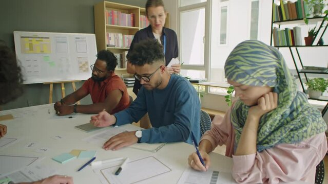 Group of multi-ethnic immigrant students sitting at table in classroom and writing on papers during exam in language school, female teacher checking them