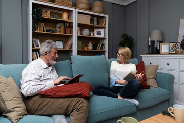 Happy senior married couple relaxing together at home with the wife sitting back from her husband as she reads a book while he surfs the internet on a tablet. Love and respect in older age concept
