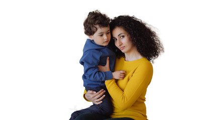 Calm Italian woman in yellow sweater, green pants sitting against transparent background, hugging son, looking at camera. Curly-haired European girl holds boy. Hispanic nanny takes care of curly child