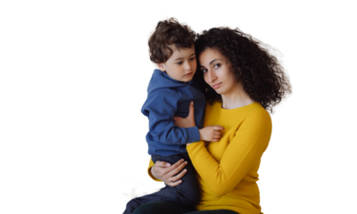 Calm Italian woman in yellow sweater, green pants sitting against transparent background, hugging son, looking at camera. Curly-haired European girl holds boy. Hispanic nanny takes care of curly child