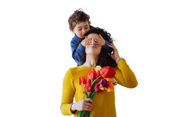 Playful hispanic curly boy stands against transparent background puts palms on mother's eyes from behind dares flowers on mothers day. Curly Italian young woman gets colourful tulips from son.