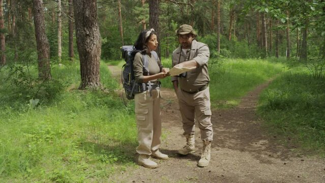 Full shot of ethnically diverse man and woman in khaki clothes and utility pants talking in forest, female tourist asking for directions and biracial male woodward showing on map and pointing to path