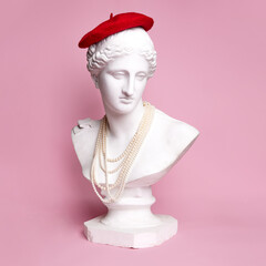 Antique statue bust of pretty woman wearing red beret and pearl necklace against pink background....
