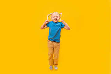 Caucasian blonde little girl with two ponytails in blue t-shirt and yellow denim. Child looking at camera and jump on yellow background. Copy space.