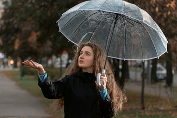 Portrait of young woman standing under transparent umbrella to see if it is raining. Girl with brown hair walks in park