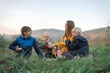 Happy mom and three kids have fun at picnic. Two boys and girl with mother drink hot drink on mountains background