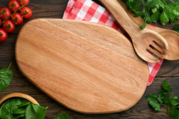 Cutting board with kitchen spoons and red napkin and parsley, tomato for cooking on old wooden dark background. Vegetarian food, health or cooking concept. Food background with free space for text.