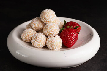 On a dark background, coconut sweets and strawberries in a light plate.