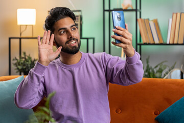 Happy indian man blogger taking selfie on smartphone, communicating video call online with social media subscribers followers, recording vlog stories. Young hindu guy at home apartment room on couch