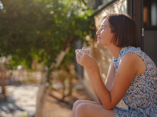 Woman is drinking coffee , morning routine near her Tiny house. First property. Small apartment in summer garden. Minimalism. Moving in. Living alone. Charming trailer house