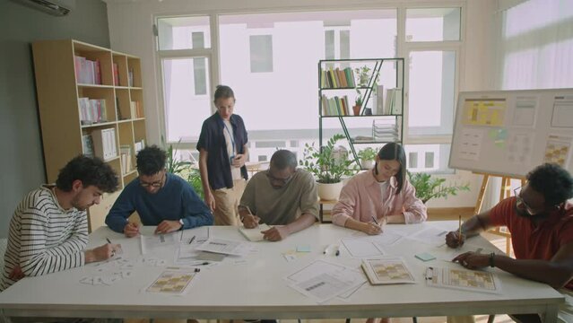 Wide shot of female teacher walking around multi-ethnic migrant students and monitoring them during test in language school