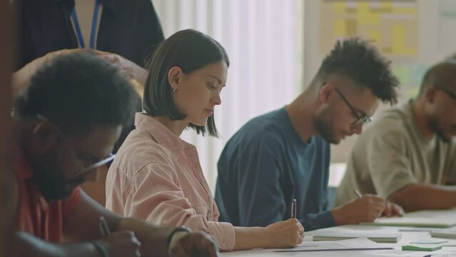 Rack focus shot of group of diverse migrant students sitting at table in classroom and writing on papers while passing exam in language school, teacher walking behind them