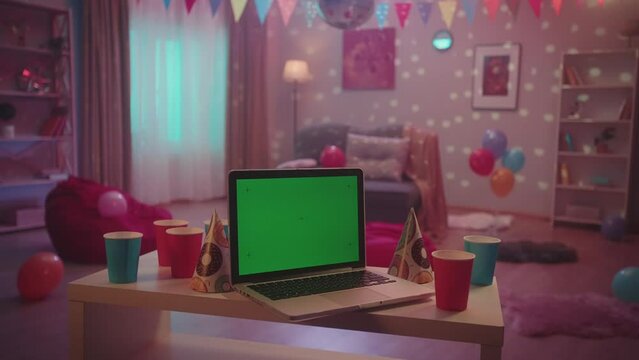 Laptop with green screen display in front of a room decorated for a house party. On a blurred background, multicolored lights flash and a disco ball sparkles. Advertising area, workspace mock up.