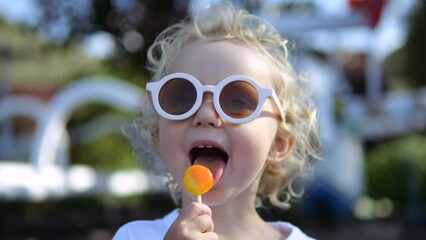 Close up portrait of kid eats caramel sweet sugary lollipop in nature park. Cute curly blonde Caucasian baby kid child is happy to eat sweet lollipop. Pleasing lollipop, capable of Generative AI