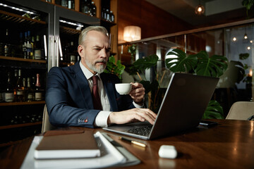 Fototapeta na wymiar Serious mature businessman working online on laptop while drinking coffee at table in cafe