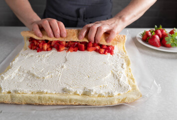 Filling a sponge cake roll with cream and strawberries and then rolls it up to a swiss roll