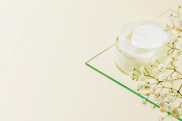 Cosmetic glass jar with natural cream on glass shelf on light beige background with white flowers. Organic beauty product and skin friendly cosmetics concept. Mockup, copy space, close up
