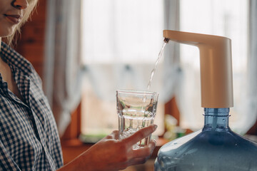 Woman's hands pour water into glass from an automatic water cooler, closeup top view. Pouring fresh clear water from blue huge bottle to glass. Clean drinking bottled water from the well.