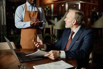 Mature businessman in suit making order to waiter while sitting at table in the restaurant during lunch time
