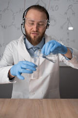 a chemistry teacher in white sits on a chair against the background of a marker board with formulas mixes two liquids conducts an experiment. online tutor concept.