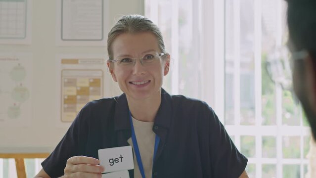 Tilt up over the shoulder shot of female teacher showing word cards with irregular verbs and talking to students during English lesson in language school