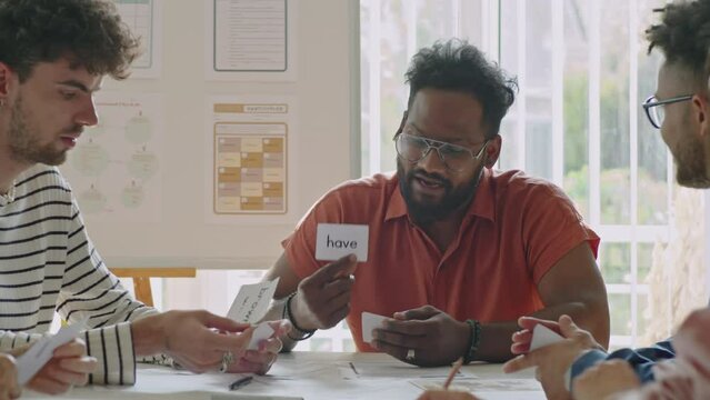 Multiracial male teacher explaining English words on cards to group of immigrant students sitting around table while giving lesson in language school
