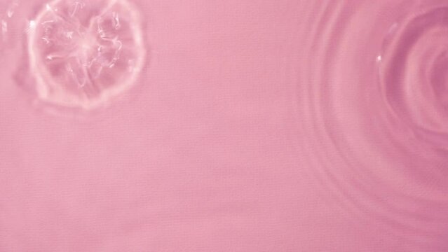 Top view slow motion closeup water surface texture with falling drops and diverging circles of water on pink background